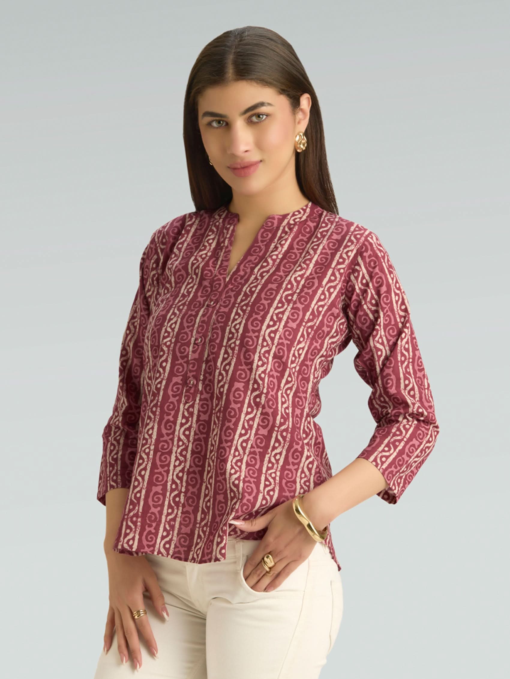 Woman wearing a maroon patterned blouse with white pants on a gray background