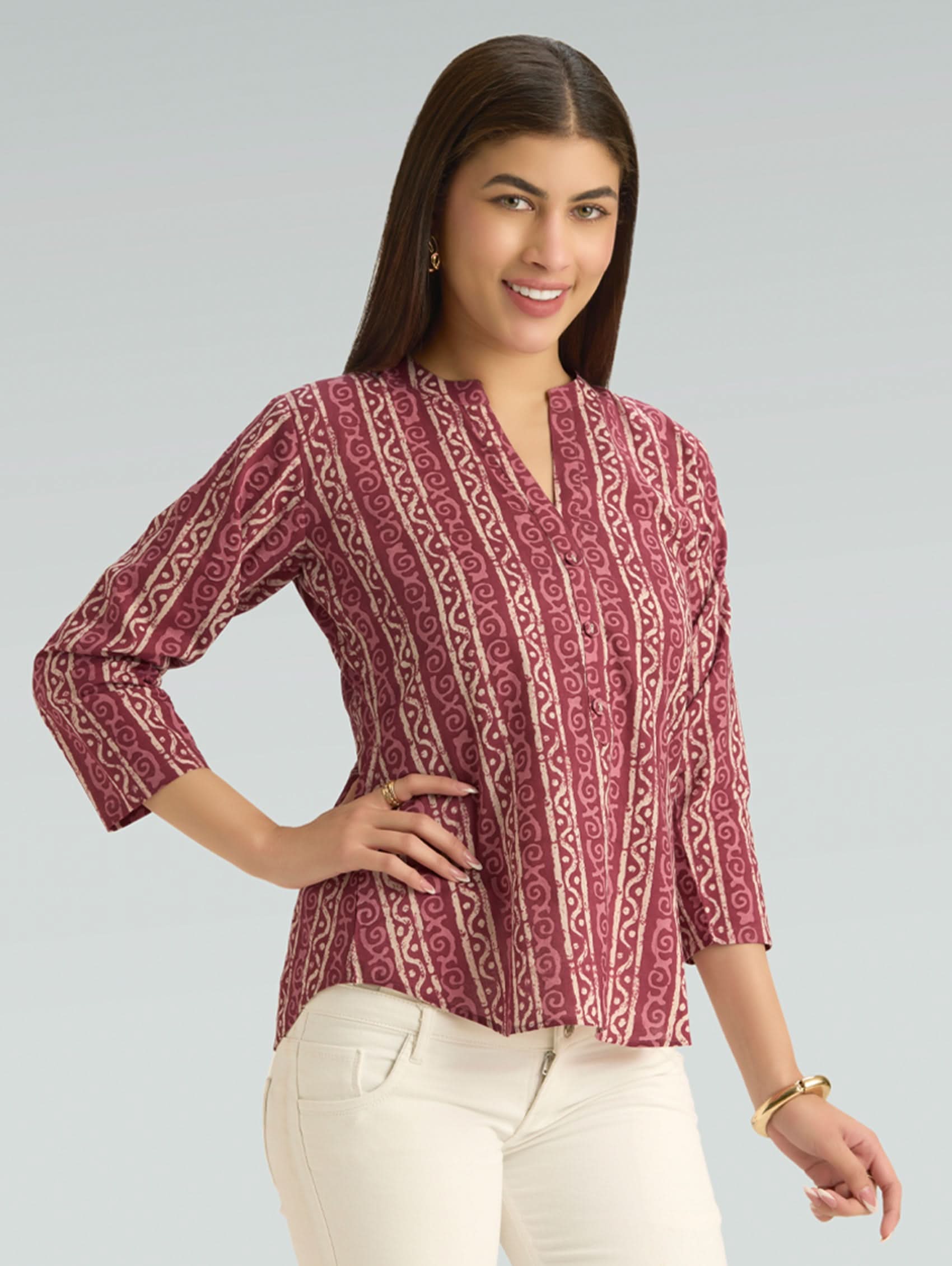 Woman wearing a patterned maroon top with white pants on a gray background
