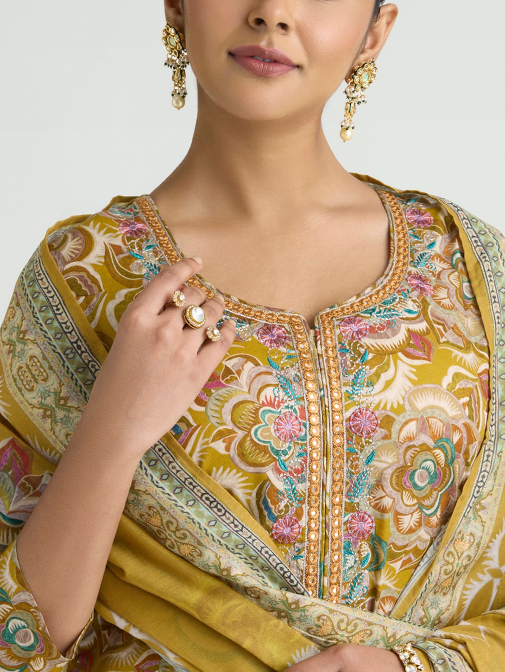 Woman wearing a traditional embroidered outfit with gold jewelry against a neutral background