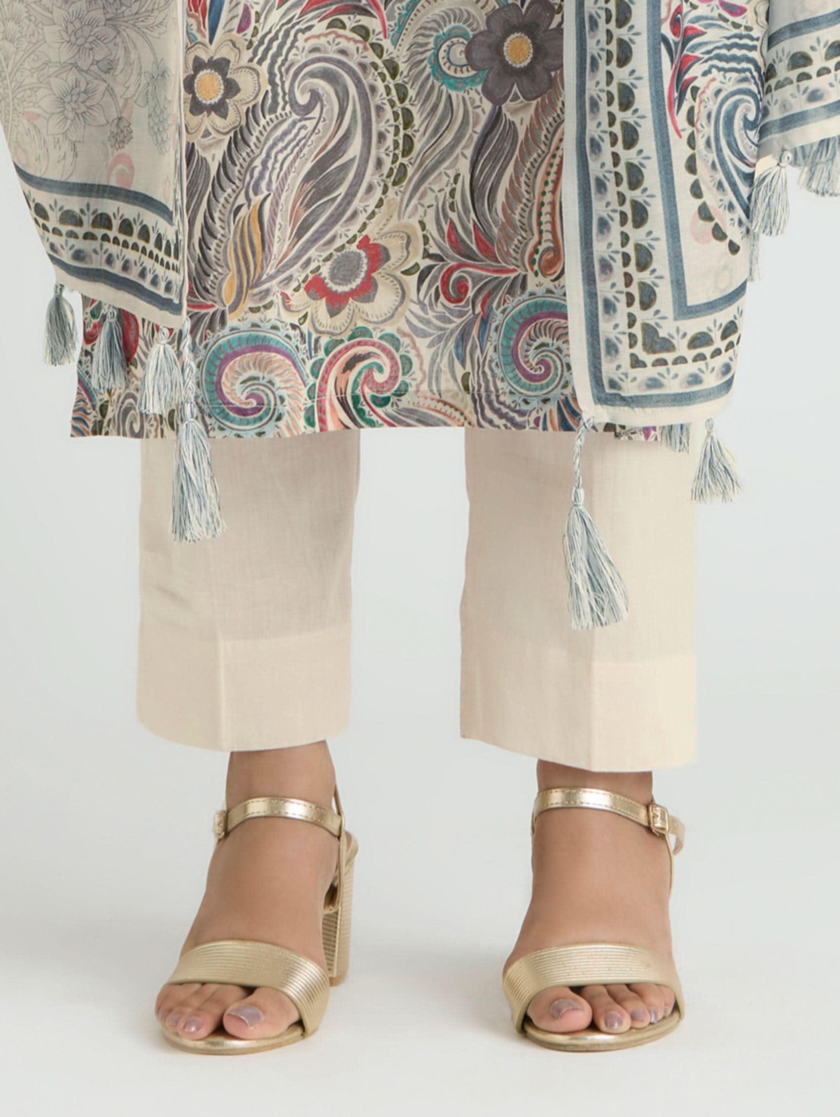 Person wearing a patterned top with tassels and beige pants, paired with gold sandals on a white background.