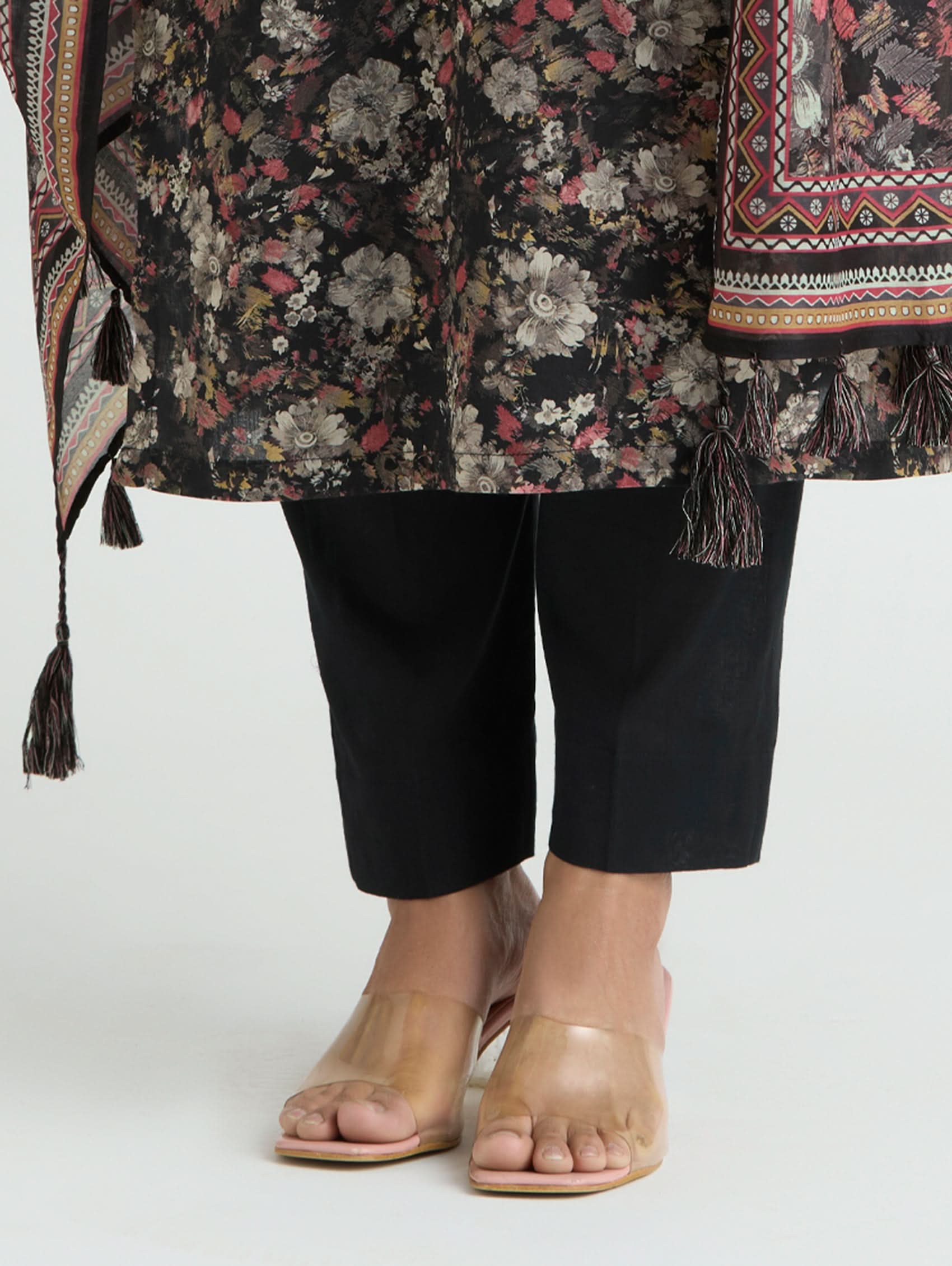 Person wearing a floral patterned coat with tassels, black pants, and beige sandals on a white background