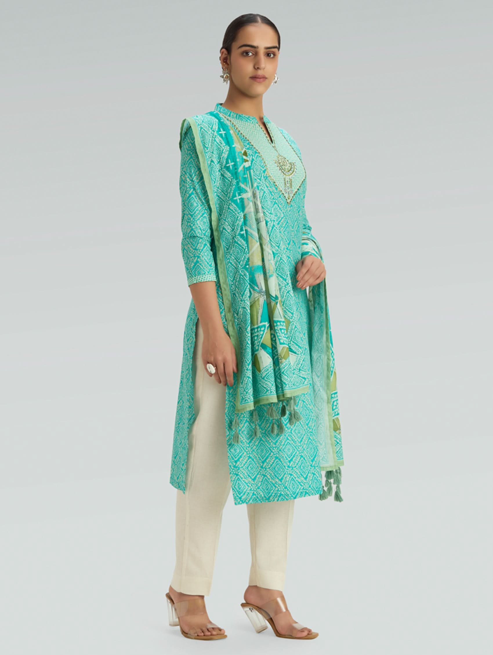 Woman wearing a turquoise embroidered outfit with white pants on a gray background