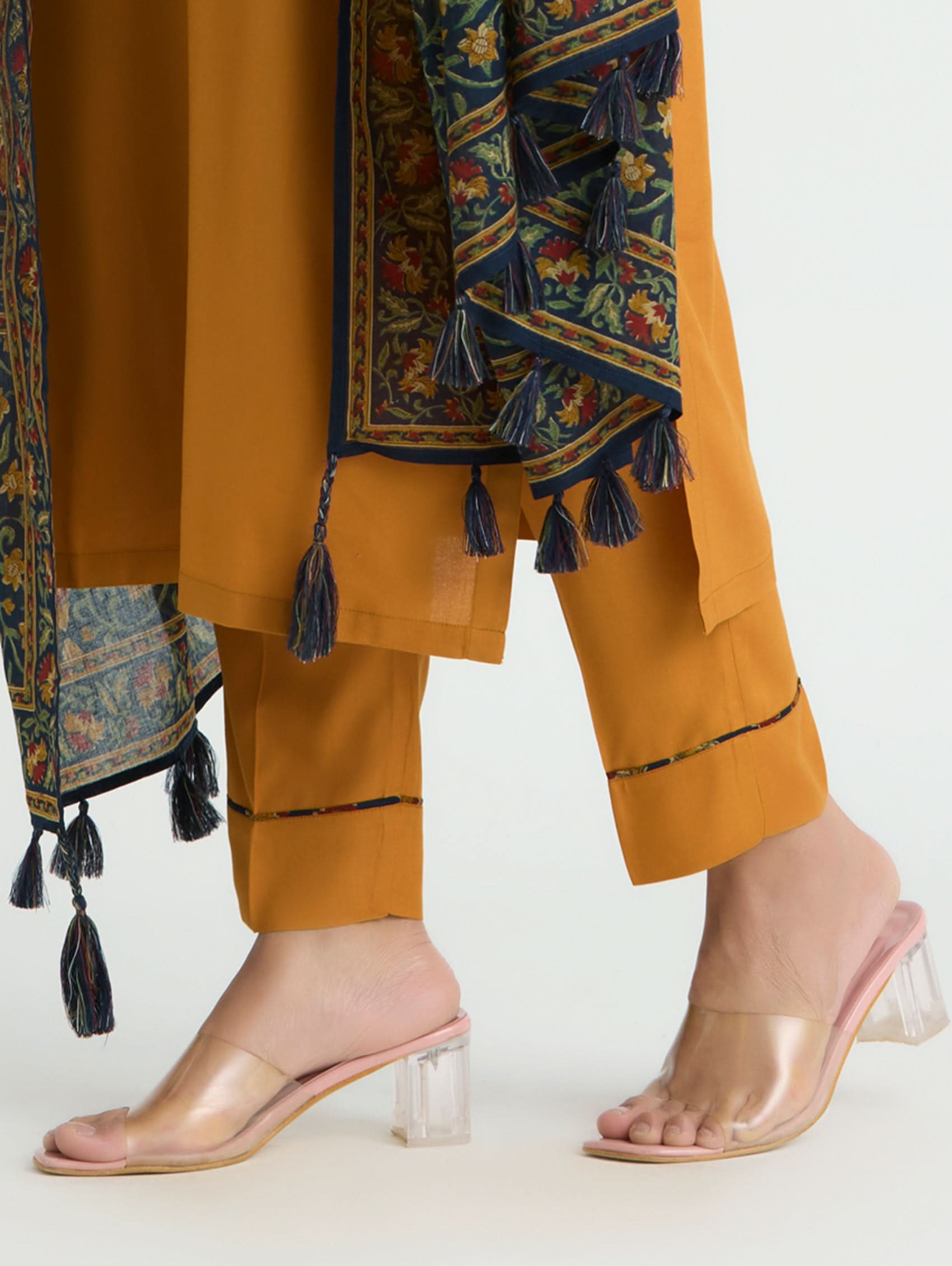 Mustard-colored pants with a patterned shawl and clear high-heeled sandals on a white background