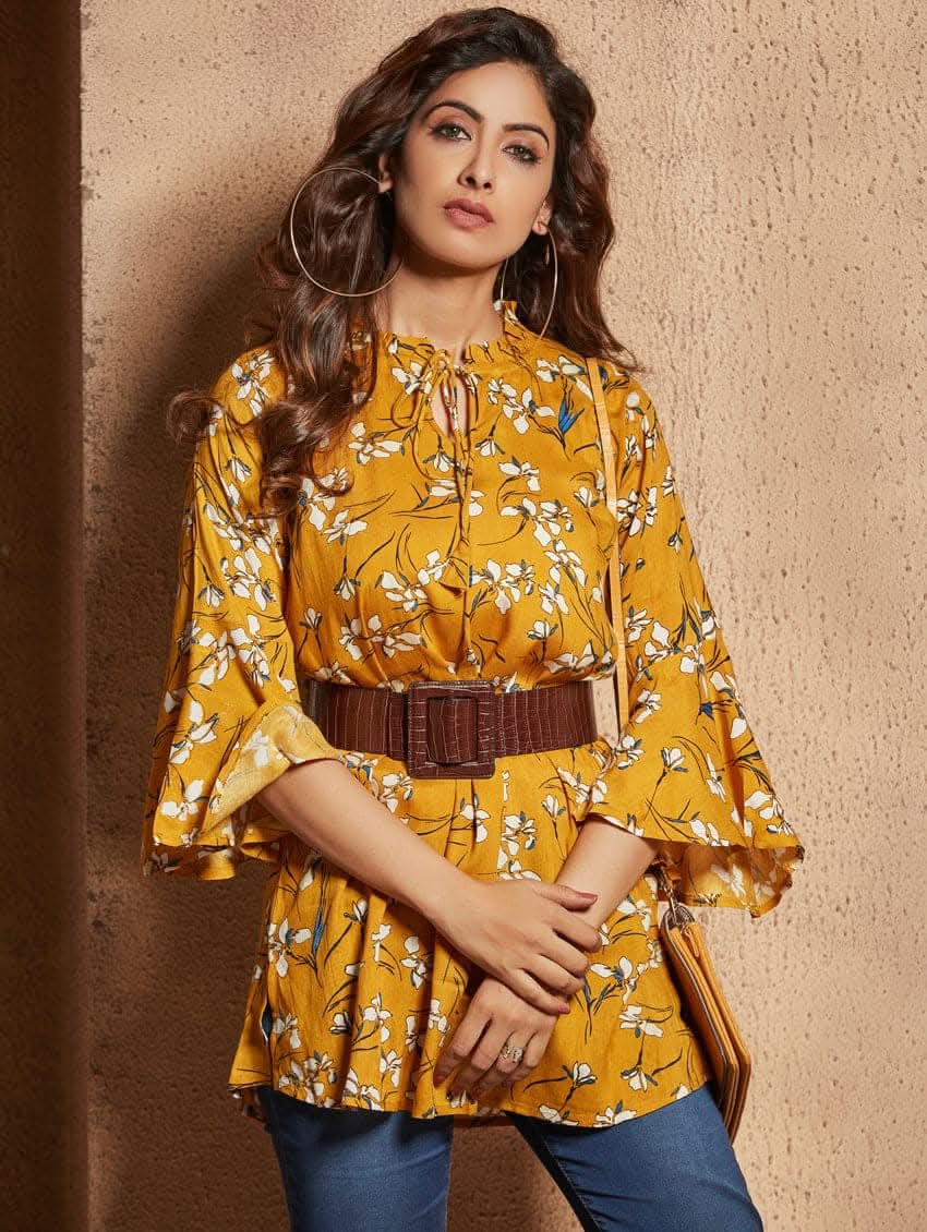 A woman wearing a mustard yellow floral print tunic with a round neckline and three-quarter bell sleeves, paired with blue jeans and tan ankle boots.