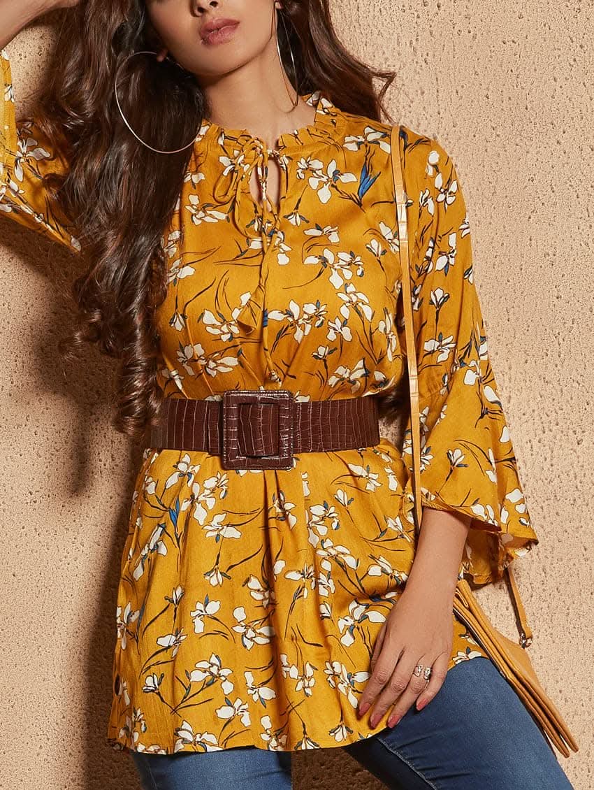 A woman wearing a mustard yellow floral print tunic with a round neckline and three-quarter bell sleeves, paired with blue jeans and tan ankle boots.