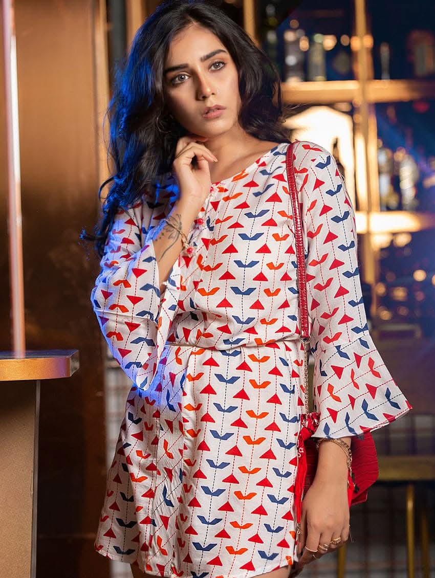 A woman wearing a printed rayon tunic with a round neckline and three-quarter bell sleeves, accessorized with a red belt and bag, standing in a bar setting.