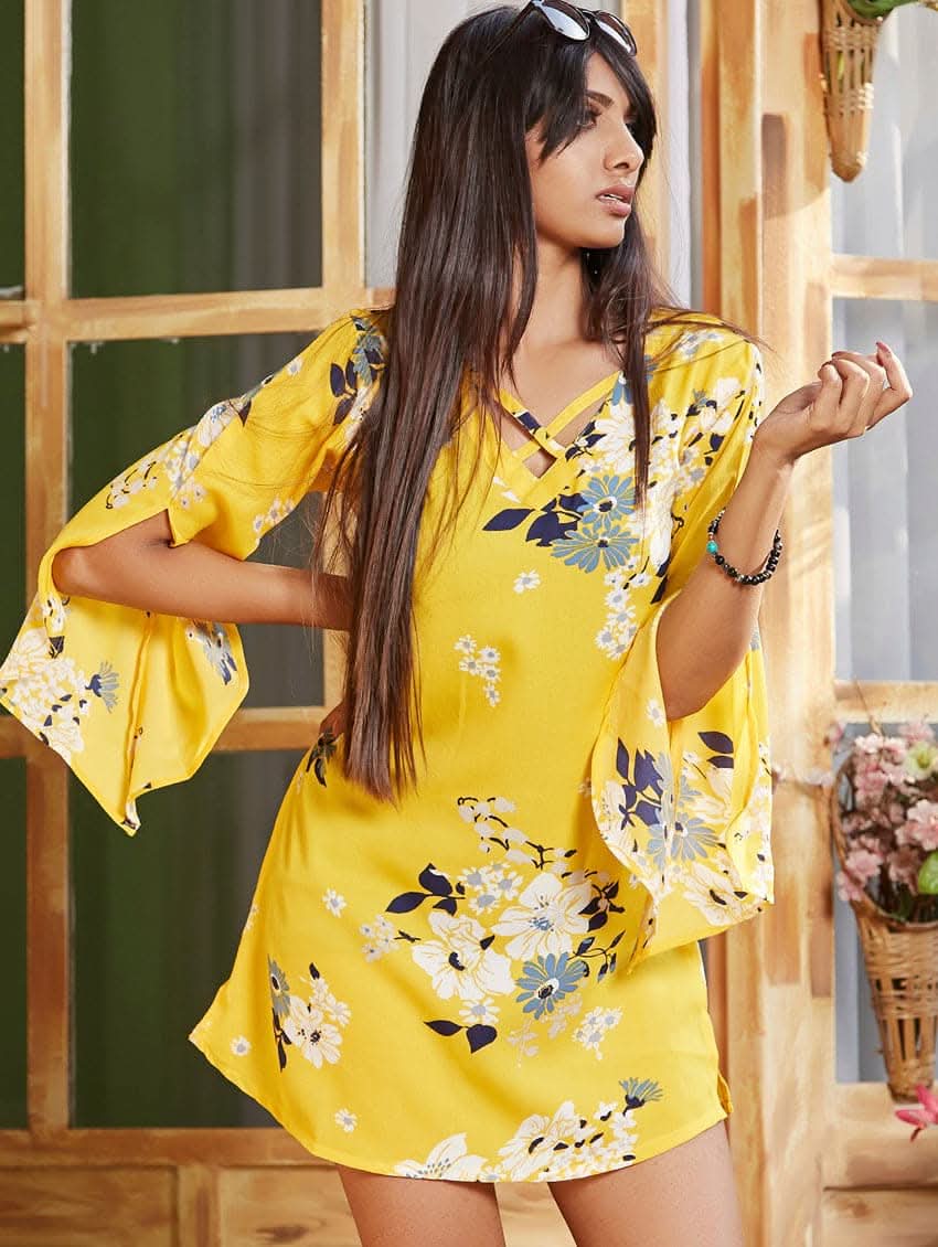 A woman wearing a yellow floral print tunic with V-neckline and three-fourth bell sleeves, standing in a doorway.