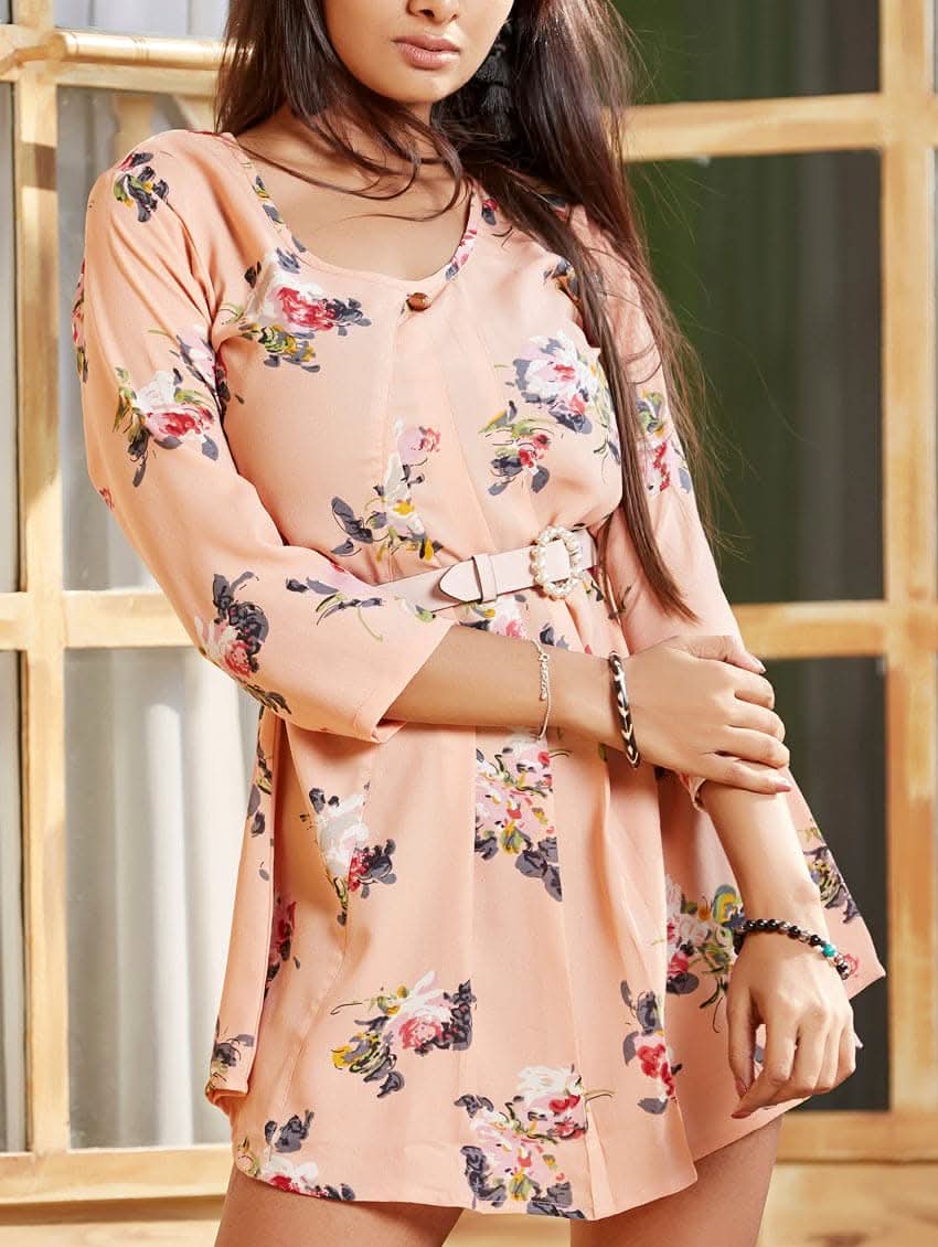 A woman wearing a beige floral print tunic with a round neckline and three-fourth bell sleeves. She is also wearing light-colored pants and sandals, posing in front of a wooden door.