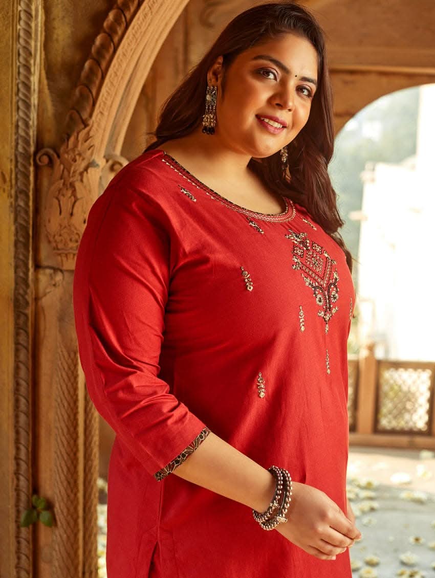 A woman standing in a beautifully decorated room wearing a red handloom cotton kurta with round neckline and three fourth sleeves, featuring computerised thread embroidery.