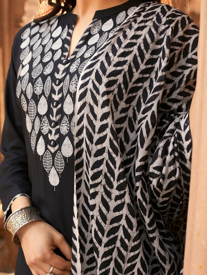 A woman wearing a black rayon suit set with embroidery on the kurta, consisting of a solid colored kurta with pants and a printed mul dupatta.