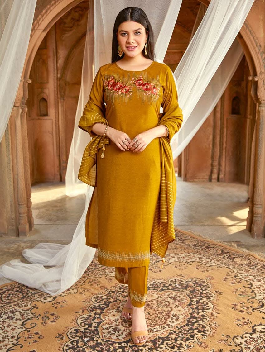 A woman wearing an embroidered silk suit set with a kurta, pants, and a dupatta, standing in a decorative setting.