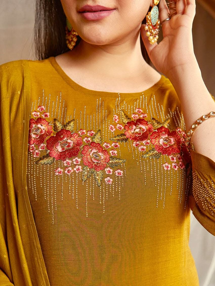 A woman wearing an embroidered silk suit set with a kurta, pants, and a dupatta, standing in a decorative setting.