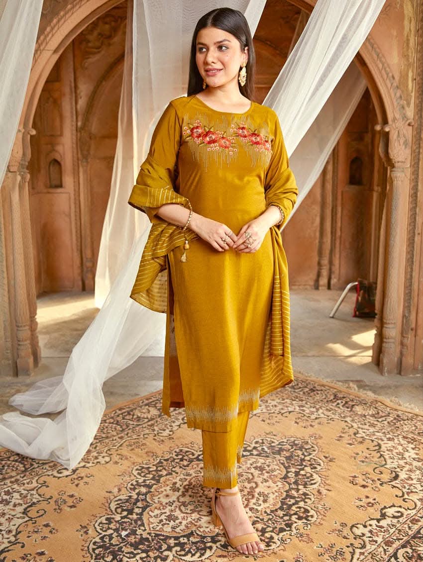 A woman wearing an embroidered silk suit set with a kurta, pants, and a dupatta, standing in a decorative setting.
