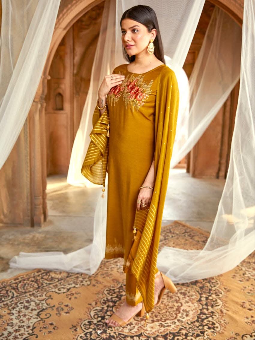 A woman wearing an embroidered silk suit set with a kurta, pants, and a dupatta, standing in a decorative setting.