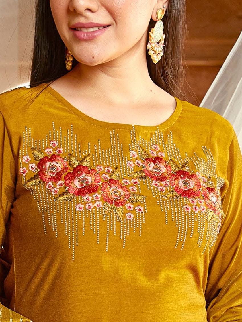 A woman wearing an embroidered silk suit set with a kurta, pants, and a dupatta, standing in a decorative setting.
