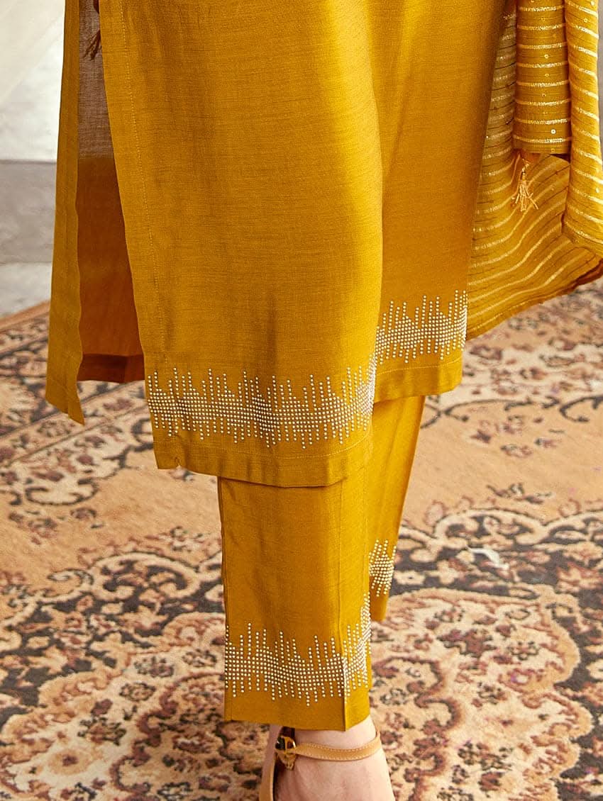 A woman wearing an embroidered silk suit set with a kurta, pants, and a dupatta, standing in a decorative setting.