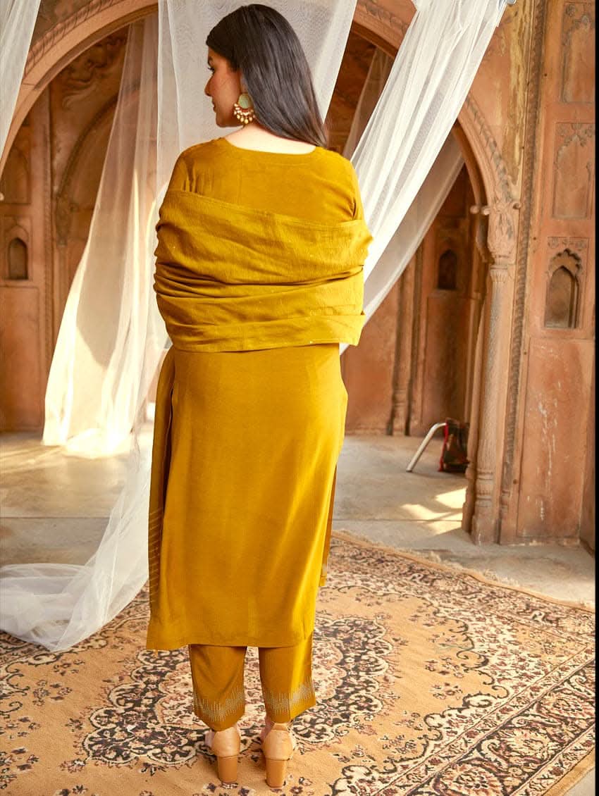 A woman wearing an embroidered silk suit set with a kurta, pants, and a dupatta, standing in a decorative setting.