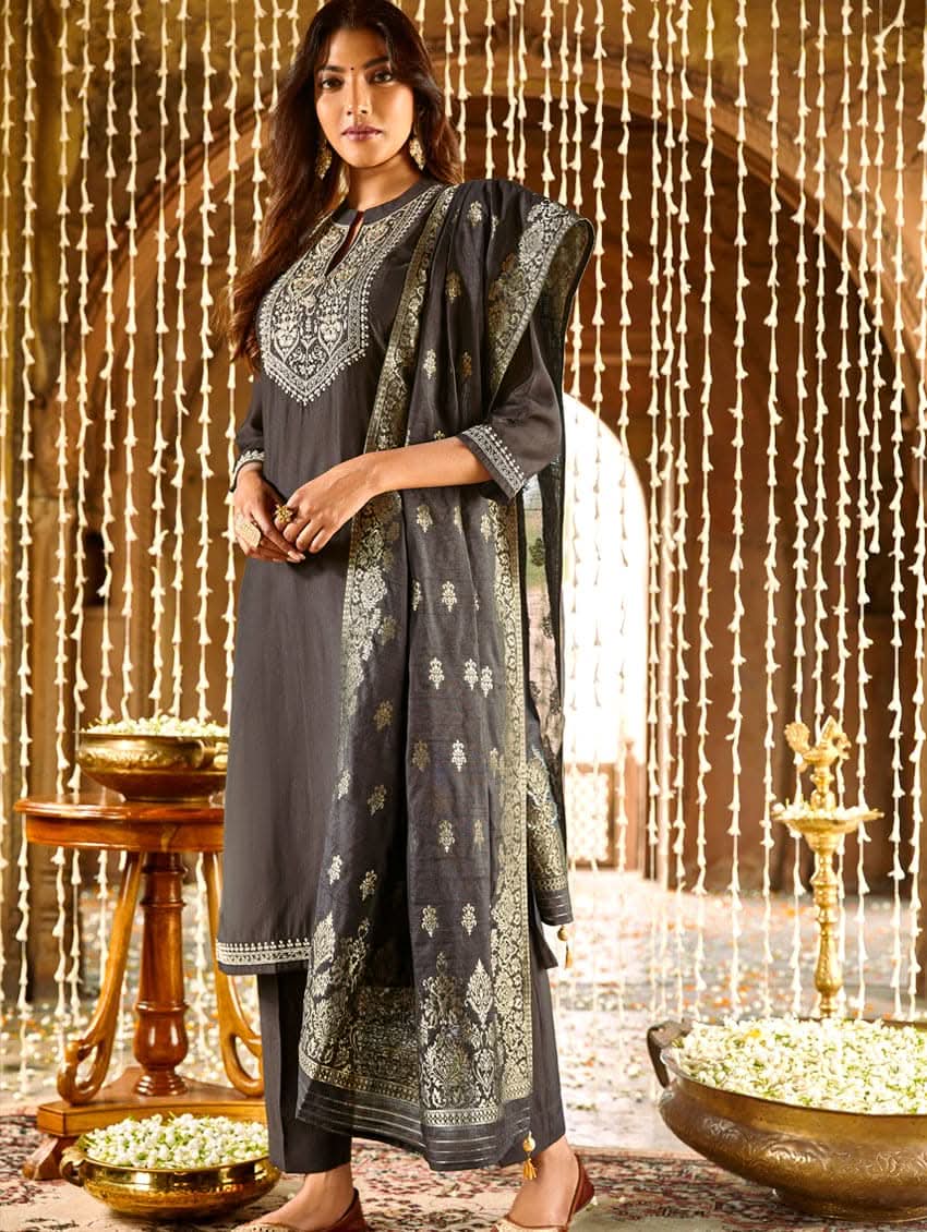 A woman modeling an embroidered silk suit set with a kurta, pants, and dupatta in a room with decorative elements.