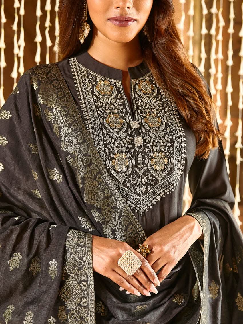 A woman modeling an embroidered silk suit set with a kurta, pants, and dupatta in a room with decorative elements.