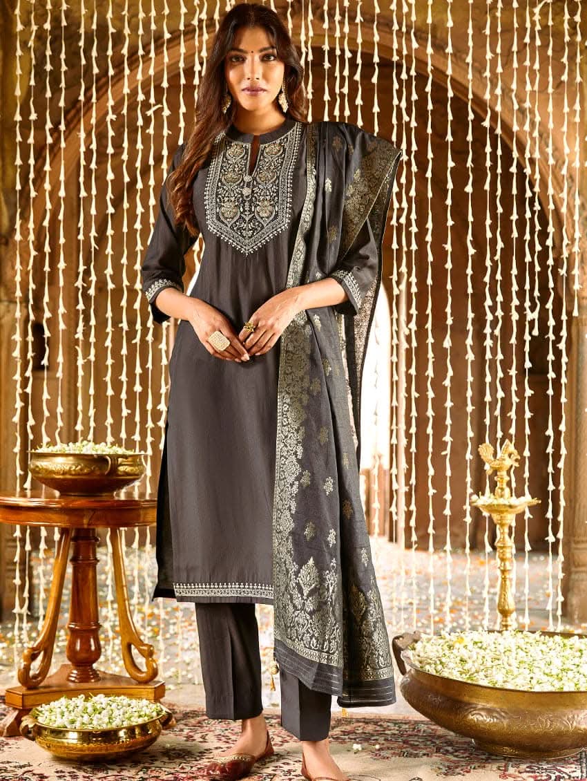 A woman modeling an embroidered silk suit set with a kurta, pants, and dupatta in a room with decorative elements.