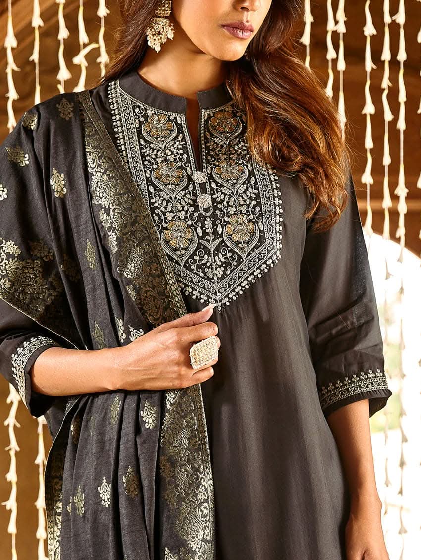 A woman modeling an embroidered silk suit set with a kurta, pants, and dupatta in a room with decorative elements.
