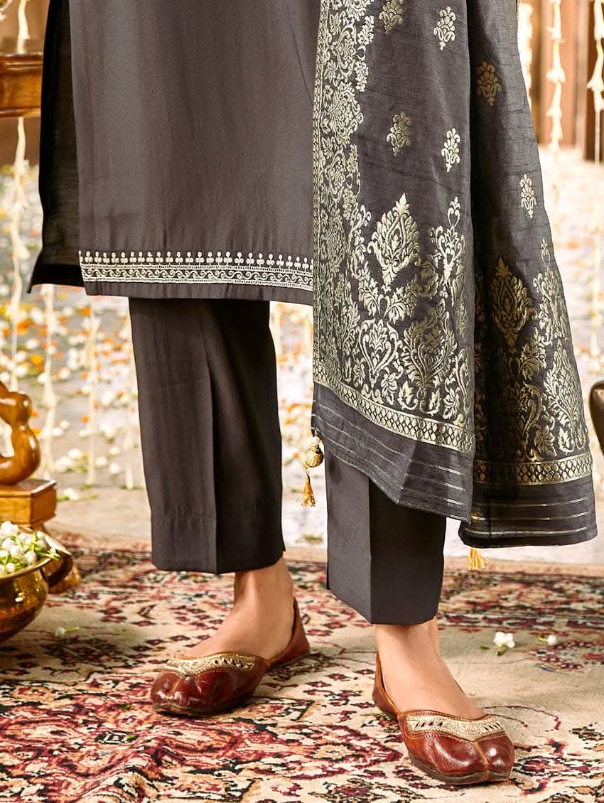 A woman modeling an embroidered silk suit set with a kurta, pants, and dupatta in a room with decorative elements.