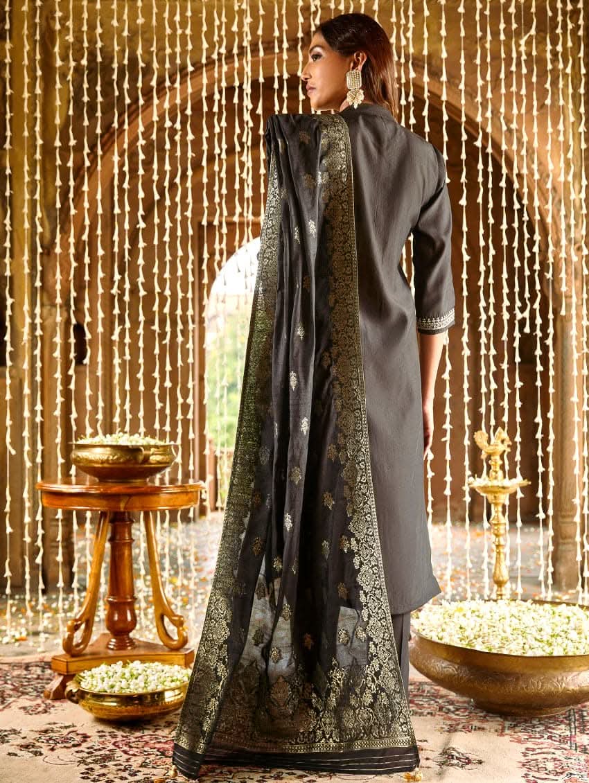 A woman modeling an embroidered silk suit set with a kurta, pants, and dupatta in a room with decorative elements.
