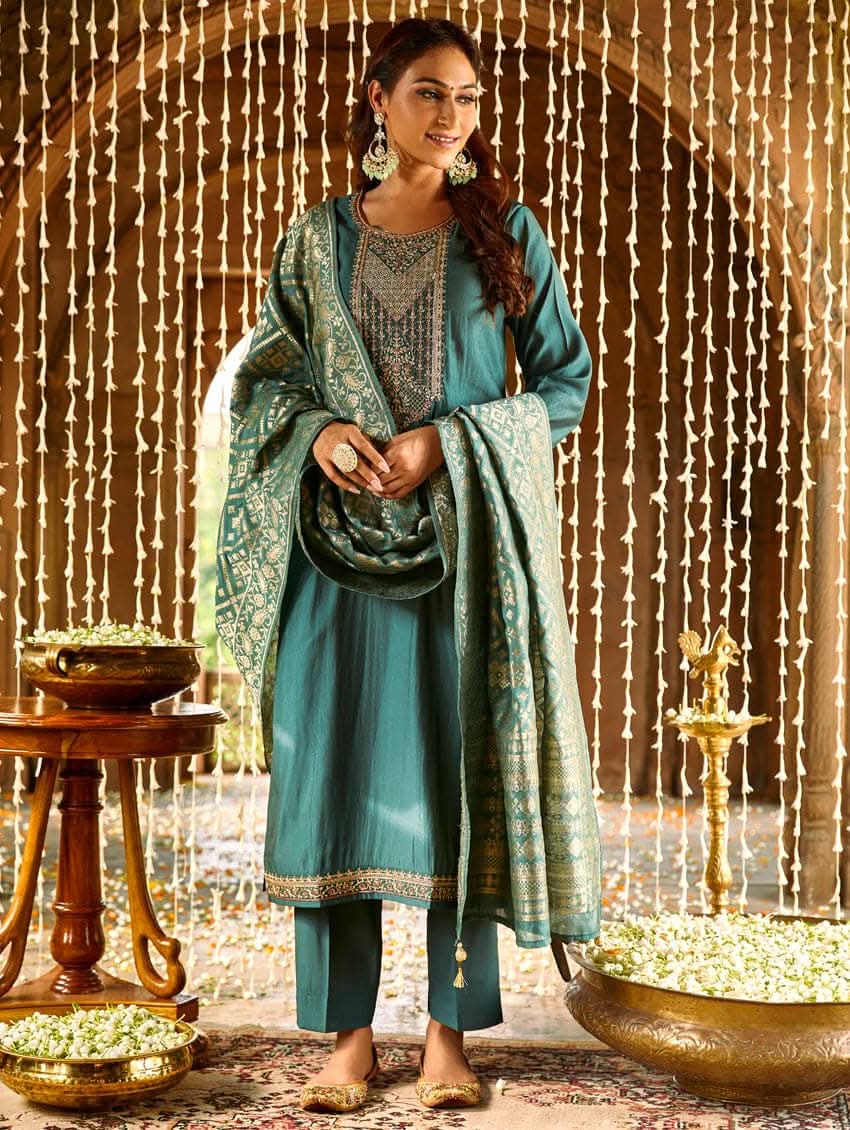 A woman standing in a beautifully decorated room, wearing an embroidered green silk suit set with a dupatta, holding a small container, with decorative elements like hanging ornaments and a patterned floor in the background.