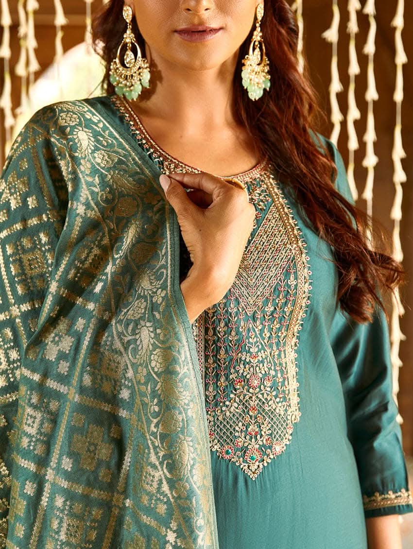 A woman standing in a beautifully decorated room, wearing an embroidered green silk suit set with a dupatta, holding a small container, with decorative elements like hanging ornaments and a patterned floor in the background.