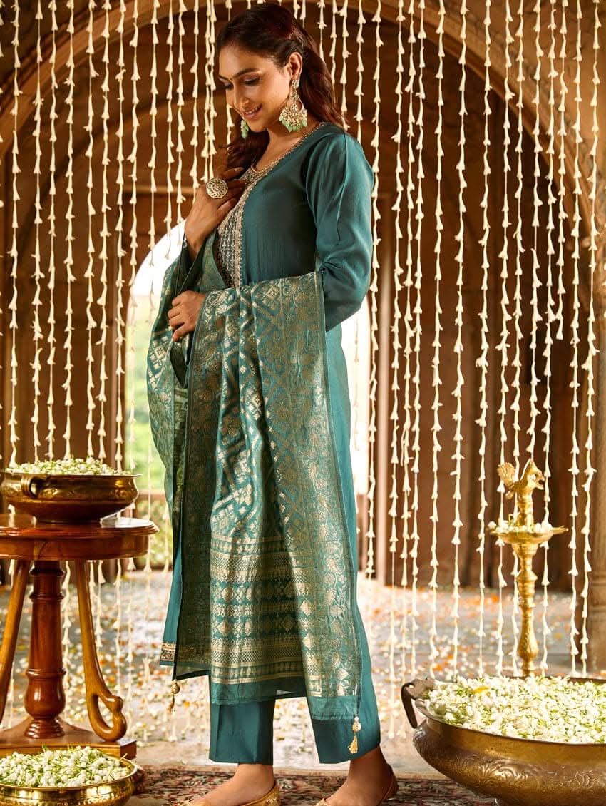 A woman standing in a beautifully decorated room, wearing an embroidered green silk suit set with a dupatta, holding a small container, with decorative elements like hanging ornaments and a patterned floor in the background.
