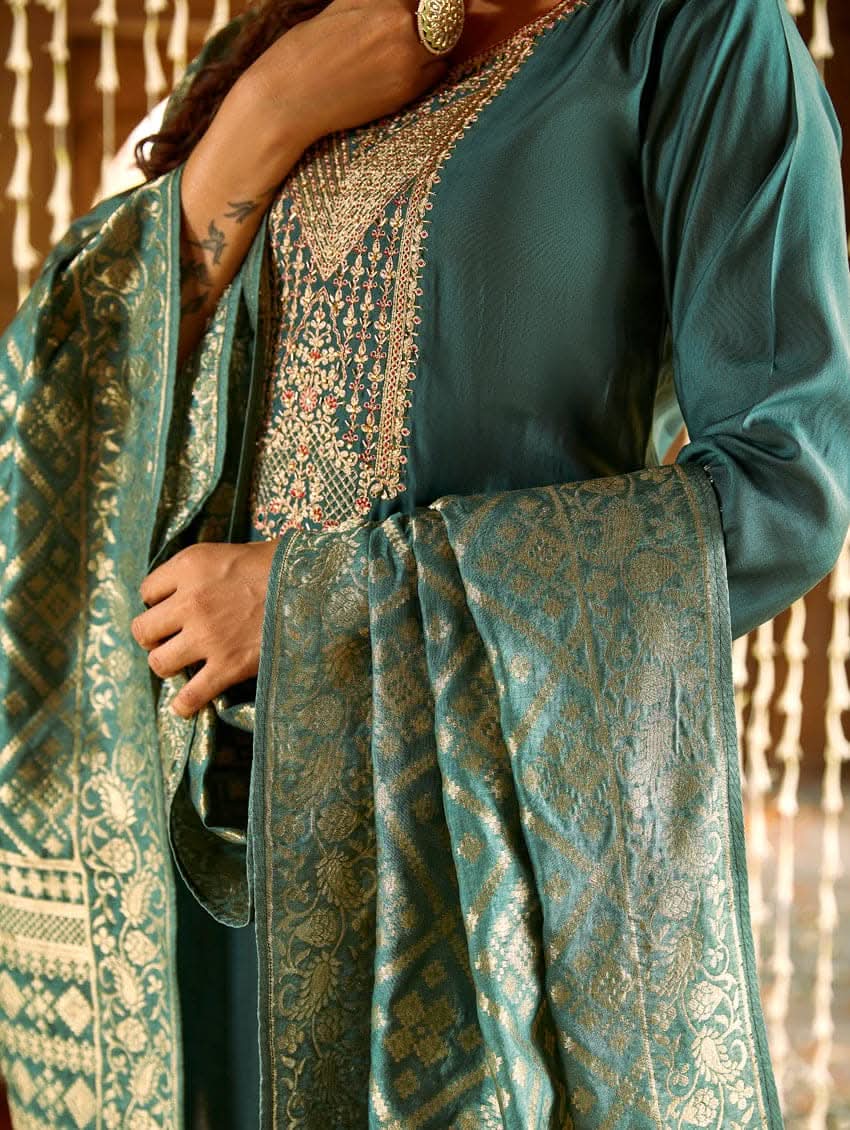 A woman standing in a beautifully decorated room, wearing an embroidered green silk suit set with a dupatta, holding a small container, with decorative elements like hanging ornaments and a patterned floor in the background.