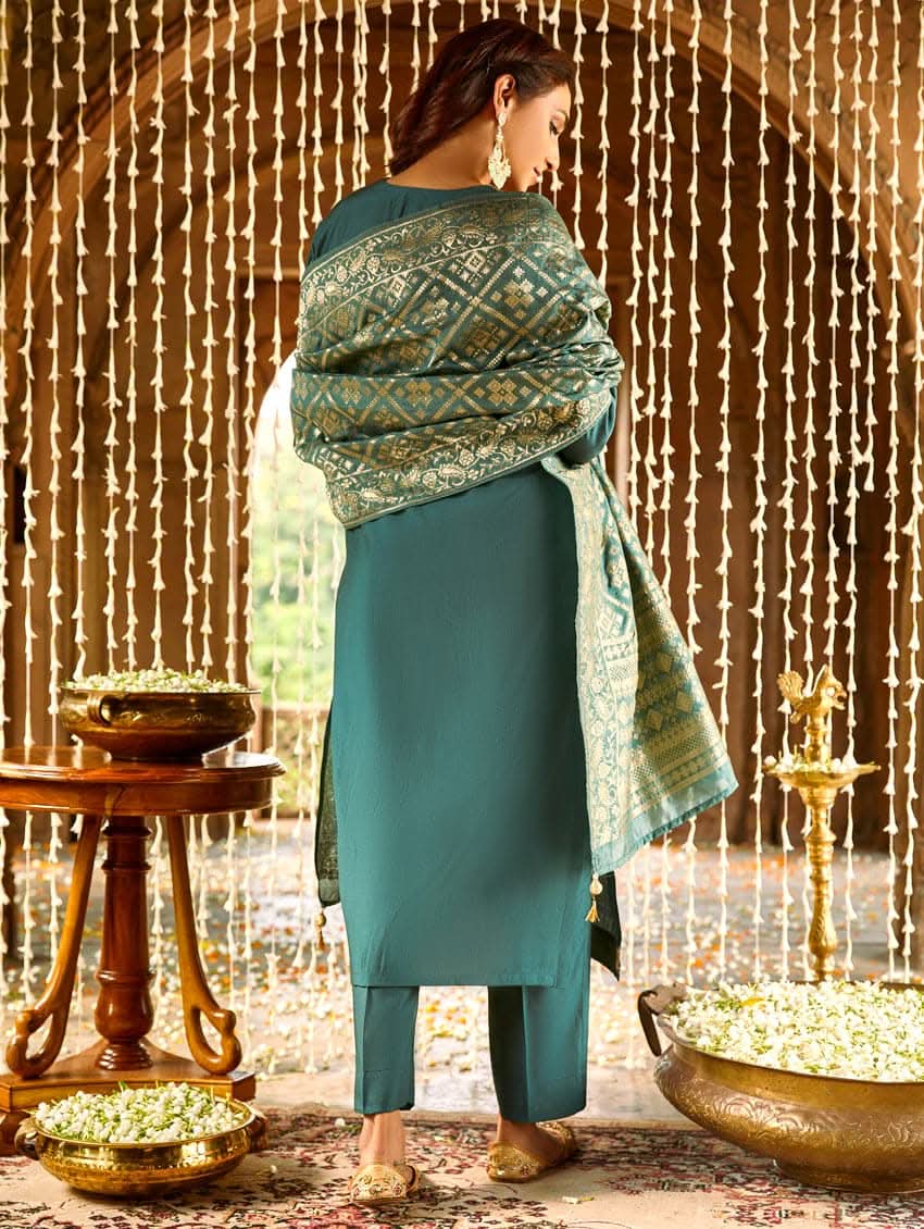 A woman standing in a beautifully decorated room, wearing an embroidered green silk suit set with a dupatta, holding a small container, with decorative elements like hanging ornaments and a patterned floor in the background.