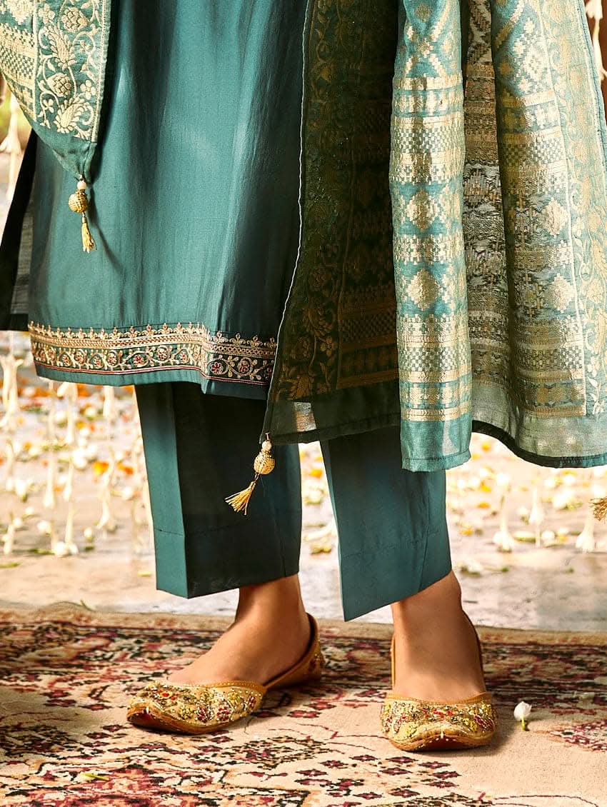 A woman standing in a beautifully decorated room, wearing an embroidered green silk suit set with a dupatta, holding a small container, with decorative elements like hanging ornaments and a patterned floor in the background.