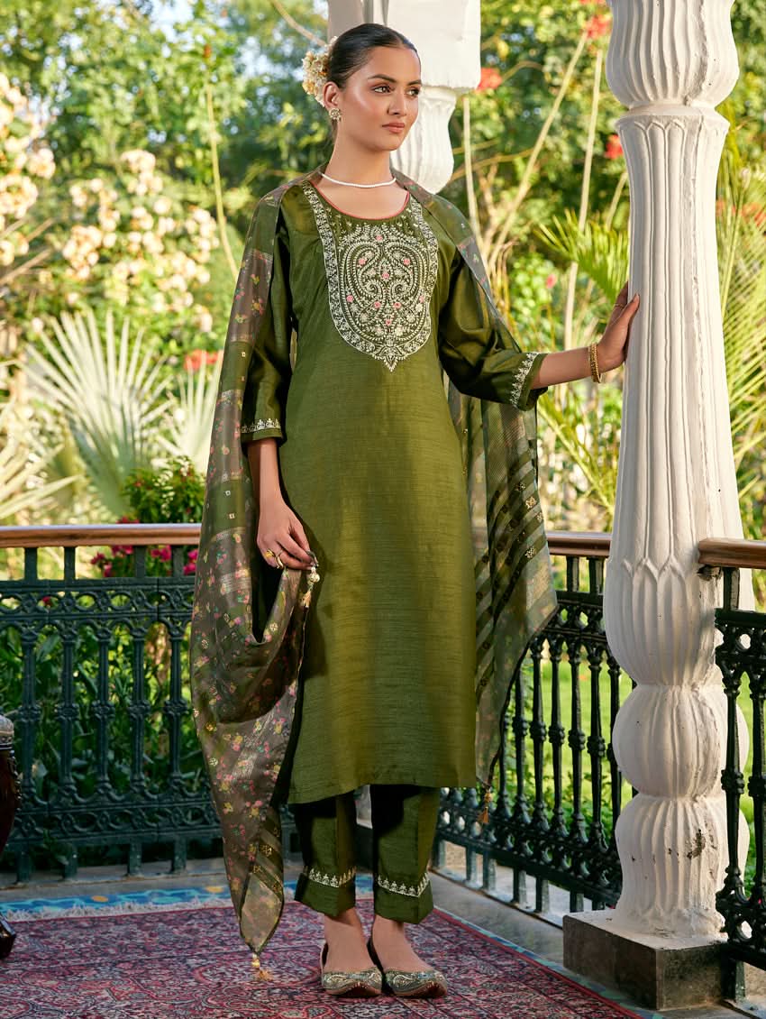 A woman standing in a columned hallway wearing a Green silk suit set with embroidered details, consisting of a kurta with round neckline and three-fourth sleeves, and pants with a partially elasticated waistband. The dupatta is also visible and printed,