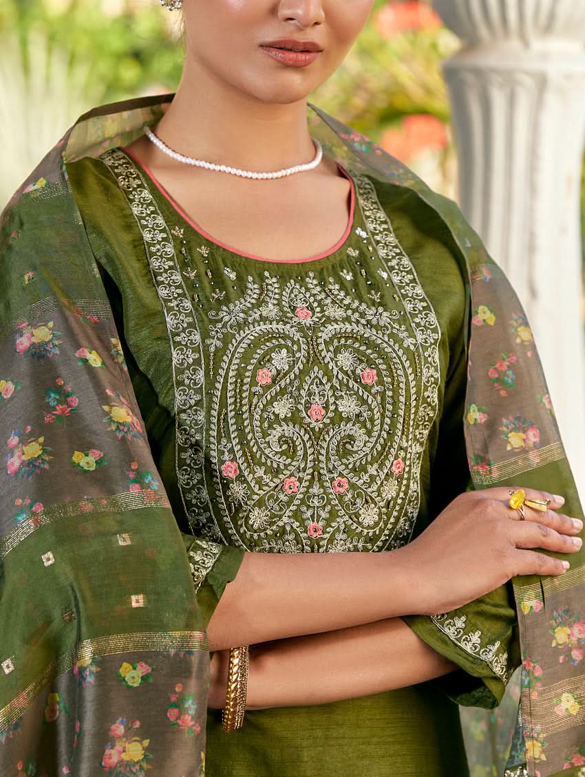 A woman standing in a columned hallway wearing a Green silk suit set with embroidered details, consisting of a kurta with round neckline and three-fourth sleeves, and pants with a partially elasticated waistband. The dupatta is also visible and printed,