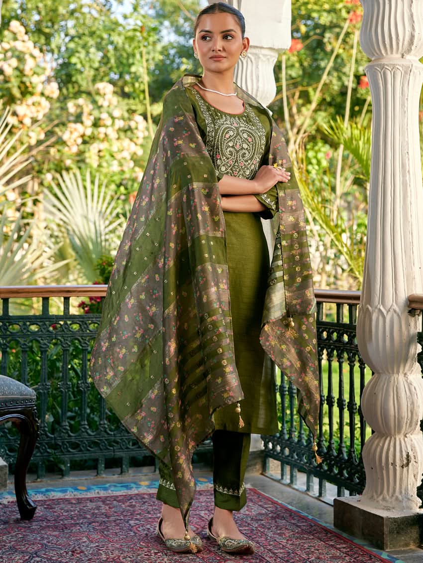 A woman standing in a columned hallway wearing a Green silk suit set with embroidered details, consisting of a kurta with round neckline and three-fourth sleeves, and pants with a partially elasticated waistband. The dupatta is also visible and printed,