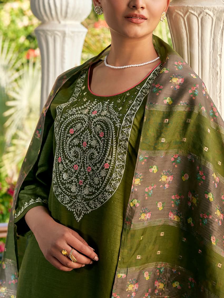 A woman standing in a columned hallway wearing a Green silk suit set with embroidered details, consisting of a kurta with round neckline and three-fourth sleeves, and pants with a partially elasticated waistband. The dupatta is also visible and printed,