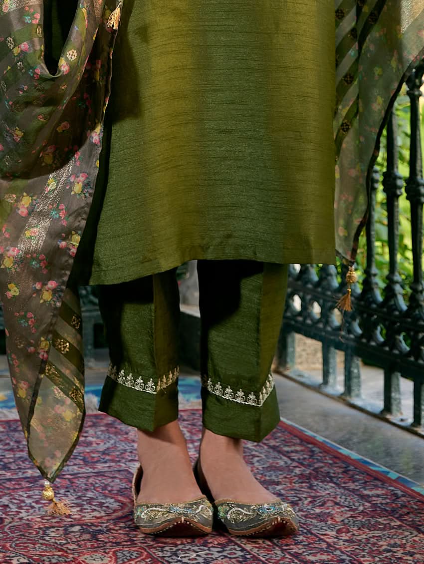 A woman standing in a columned hallway wearing a Green silk suit set with embroidered details, consisting of a kurta with round neckline and three-fourth sleeves, and pants with a partially elasticated waistband. The dupatta is also visible and printed,