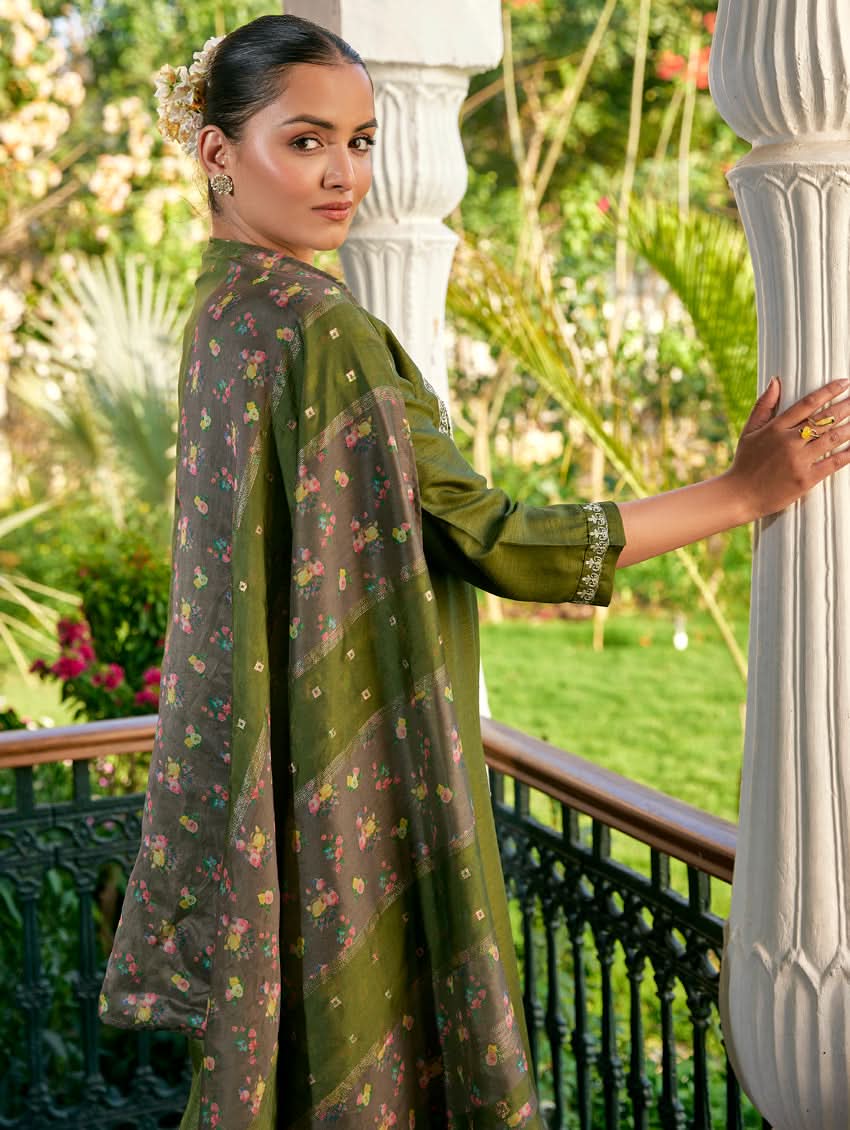 A woman standing in a columned hallway wearing a Green silk suit set with embroidered details, consisting of a kurta with round neckline and three-fourth sleeves, and pants with a partially elasticated waistband. The dupatta is also visible and printed,