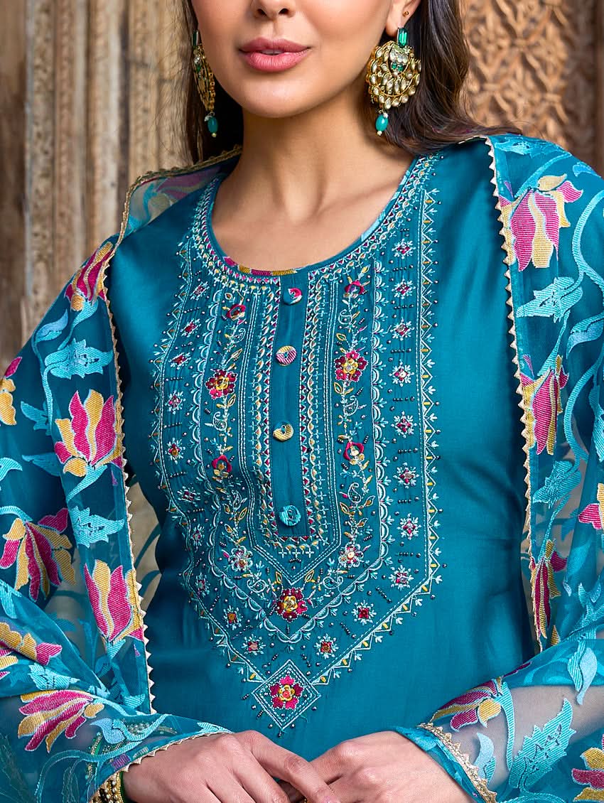 A woman standing in a beautifully decorated room, wearing a blue traditional suit with embroidery, paired with a dupatta.