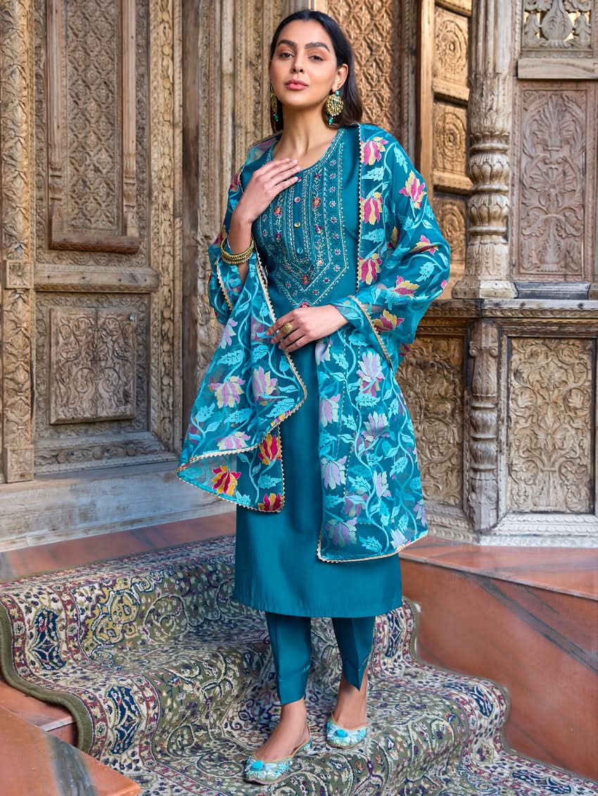 A woman standing in a beautifully decorated room, wearing a blue traditional suit with embroidery, paired with a dupatta.