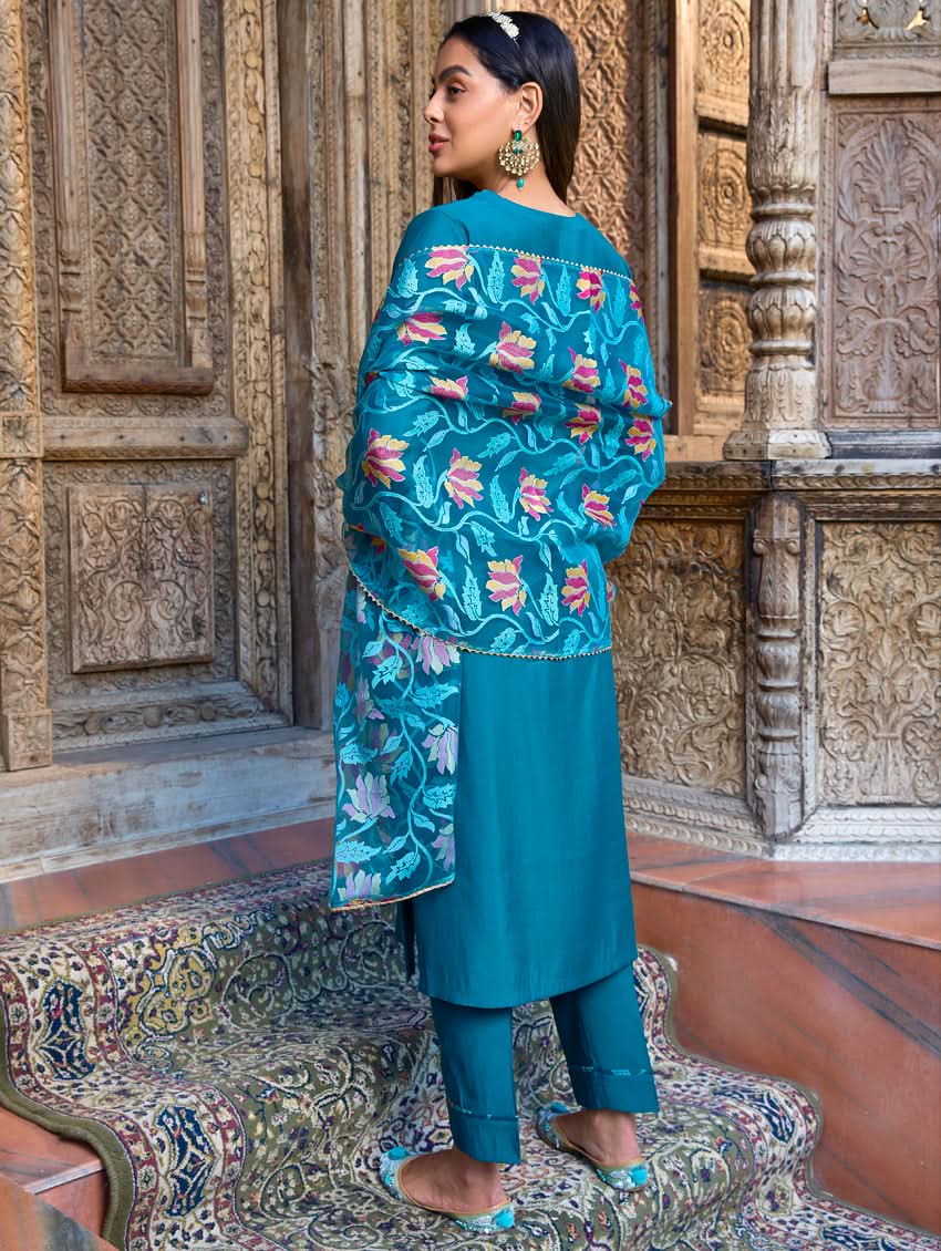 A woman standing in a beautifully decorated room, wearing a blue traditional suit with embroidery, paired with a dupatta.
