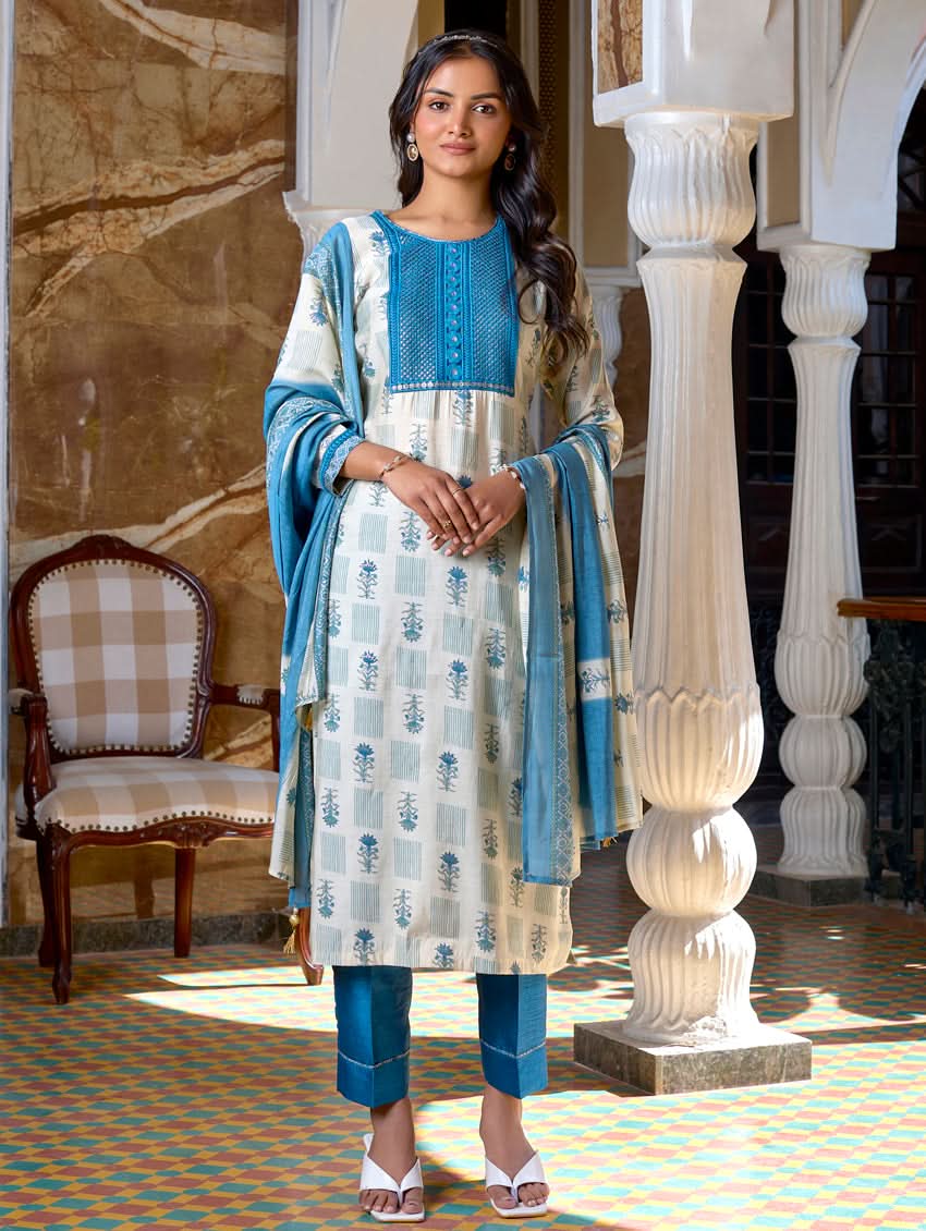 A woman standing in a beautifully decorated room, wearing a blue and white traditional suit with embroidery, paired with a dupatta.
