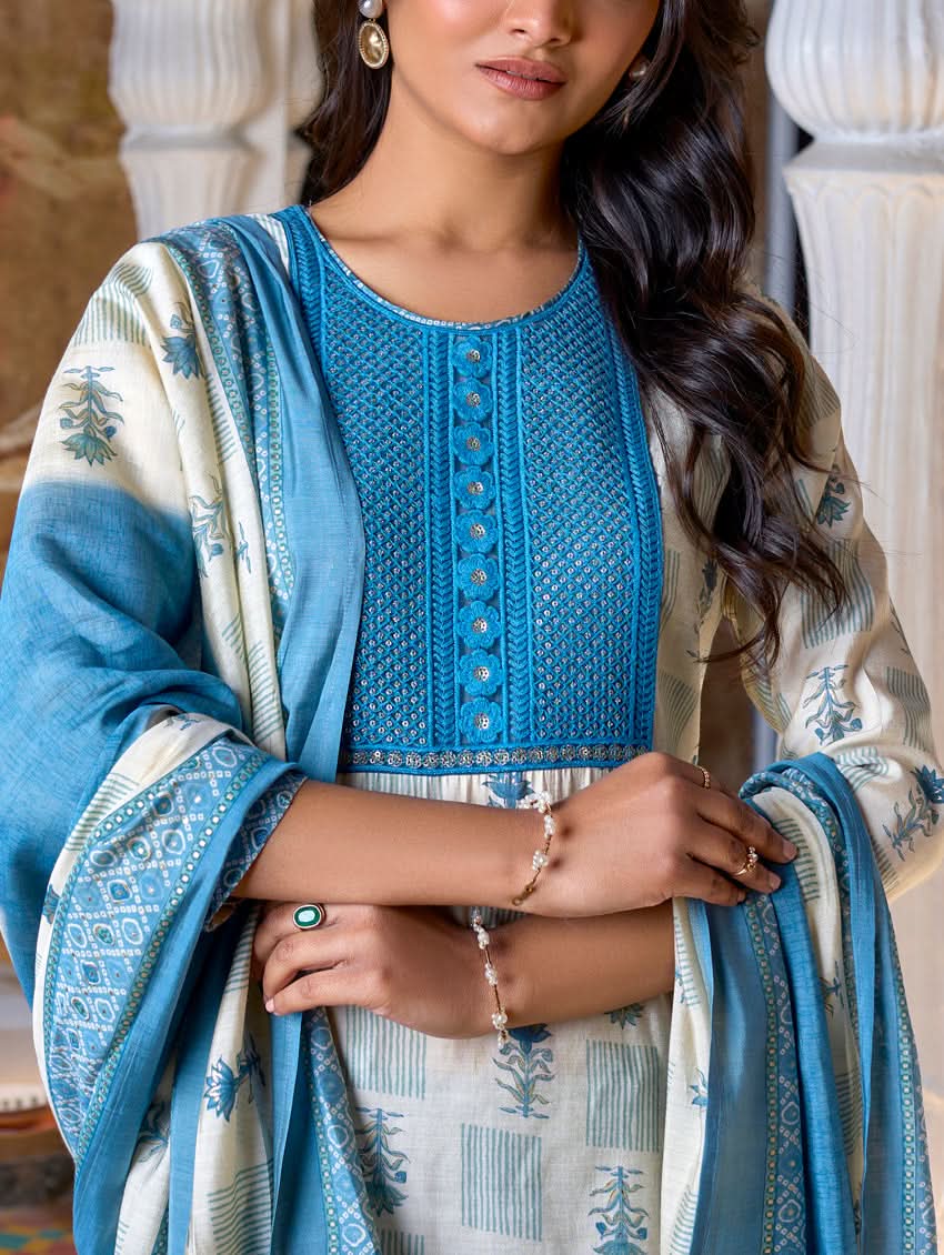 A woman standing in a beautifully decorated room, wearing a blue and white traditional suit with embroidery, paired with a dupatta.