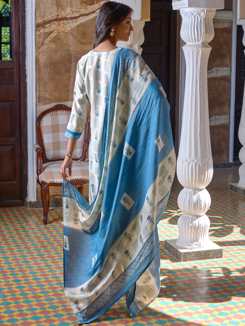 A woman standing in a beautifully decorated room, wearing a blue and white traditional suit with embroidery, paired with a dupatta.