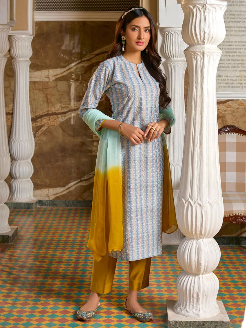 A woman wearing a traditional Schiffli embroidered silk suit set with a kurta, pants, and dupatta in a multicolored floral design, standing in a decorative room.