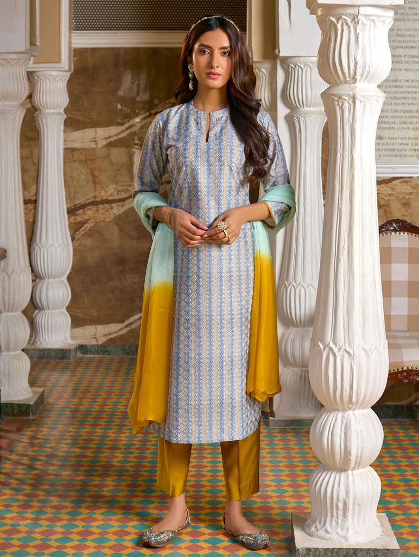 A woman wearing a traditional Schiffli embroidered silk suit set with a kurta, pants, and dupatta in a multicolored floral design, standing in a decorative room.