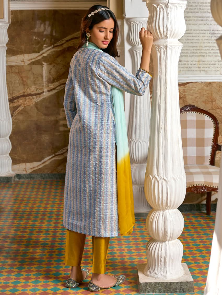 A woman wearing a traditional Schiffli embroidered silk suit set with a kurta, pants, and dupatta in a multicolored floral design, standing in a decorative room.