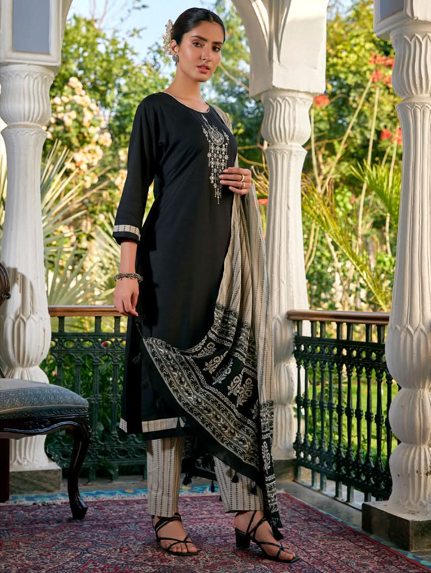 A woman standing in a columned hallway wearing a Black silk suit set with embroidered details, consisting of a kurta with round neckline and three-fourth sleeves, and pants with a partially elasticated waistband. The dupatta is also visible and printed,