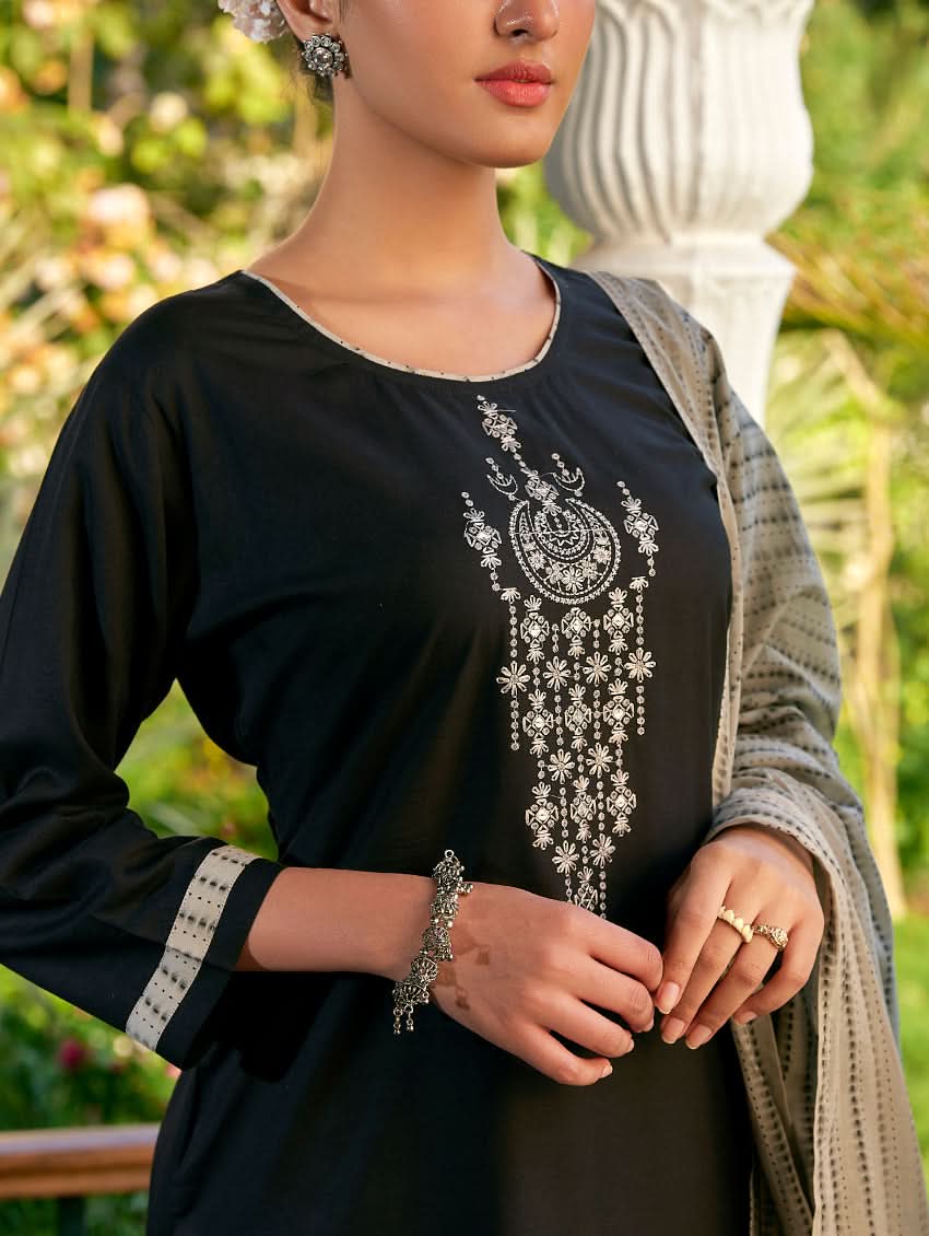 A woman standing in a columned hallway wearing a Black silk suit set with embroidered details, consisting of a kurta with round neckline and three-fourth sleeves, and pants with a partially elasticated waistband. The dupatta is also visible and printed,